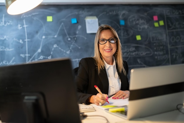 Portrait de femme chef d'entreprise au bureau du soir. Femme souriante dans des verres travaillant à table avec des ordinateurs et des papiers vérifiant les données financières en regardant la caméra. Concept de stratégie de croissance d'entreprise