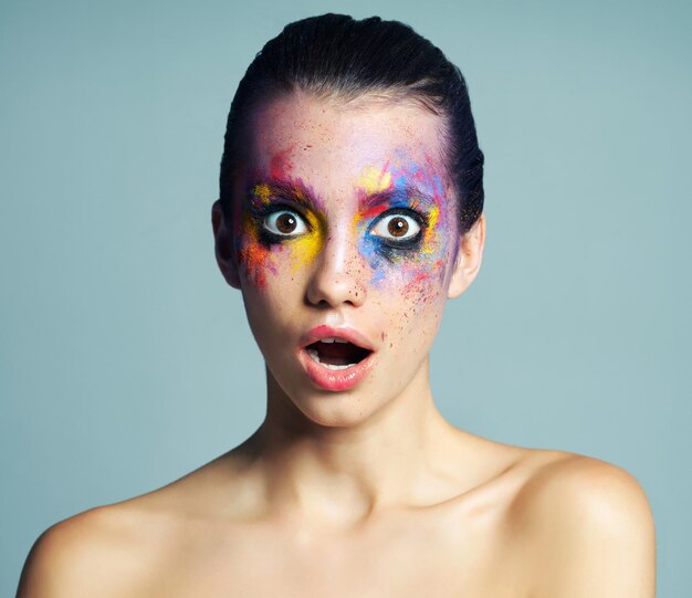 Portrait de femme et choqué avec de la peinture pour le maquillage art créatif et des cosmétiques uniques de l'esthétique Surprise fille et produit facial avec des éclaboussures de couleur pour la beauté du carnaval arc-en-ciel ou fond gris