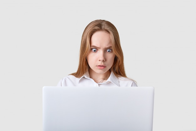 Portrait de femme choquée assise devant un ordinateur portable blanc avec les yeux grands ouverts