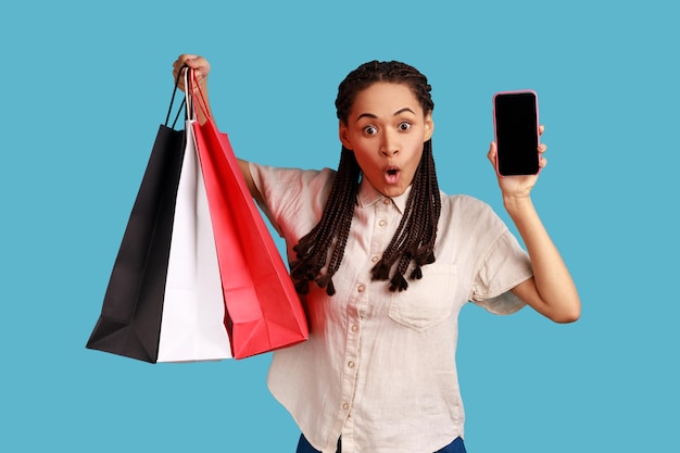 Portrait d'une femme choquée avec des dreadlocks noirs tenant des sacs à provisions et montrant un téléphone intelligent avec un écran vide pour la publicité portant une chemise blanche Prise de vue en studio intérieure isolée sur fond bleu