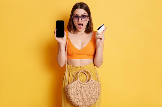 Portrait d'une femme choquée portant des vêtements d'été avec un sac de paille debout isolé sur fond jaune montrant un téléphone portable avec écran vide payant pour l'achat avec l'application bancaire