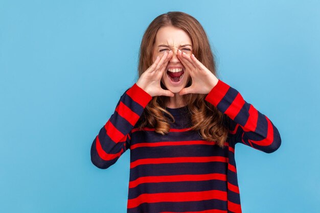 Portrait d'une femme en colère ouvre largement la bouche, crie dans la panique, découvre des nouvelles d'horreur, étant dans une situation stressante, portant un pull rayé de style décontracté Tourné en studio intérieur isolé sur fond bleu.