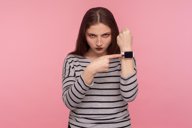 Portrait d'une femme en colère en sweat-shirt rayé nerveux à propos de l'heure tardive pointant une montre-bracelet avec affichage simulé à l'air anxieux inquiet à propos de la date limite prise de vue en studio intérieur fond rose