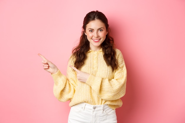 Portrait de femme confiante souriante pointant les doigts à gauche au logo, montrant un texte promotionnel avec un visage déterminé, debout contre le mur rose.