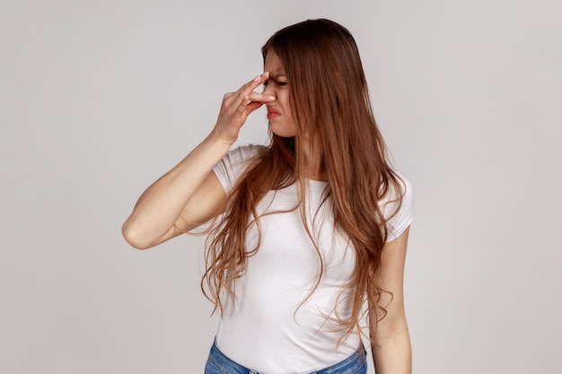 Portrait d'une femme confuse mécontente avec des cheveux noirs pinçant le nez pour éviter la mauvaise odeur exprimant la répulsion et le dégoût portant un t-shirt blanc Studio tourné en intérieur isolé sur fond gris