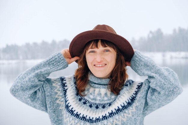 Portrait femme dans un chapeau marron et un pull sur fond de forêt et de lac enneigés