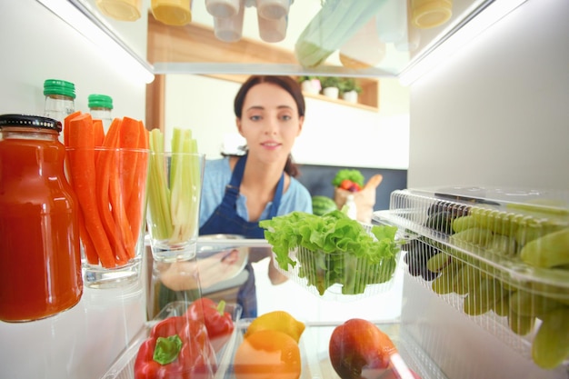 Portrait de femme debout près d'un réfrigérateur ouvert plein de légumes et de fruits sains