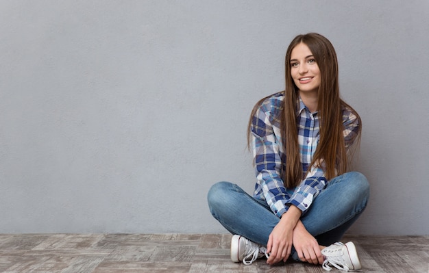 Portrait d'une femme décontractée assise sur le sol et regardant loin sur un mur gris