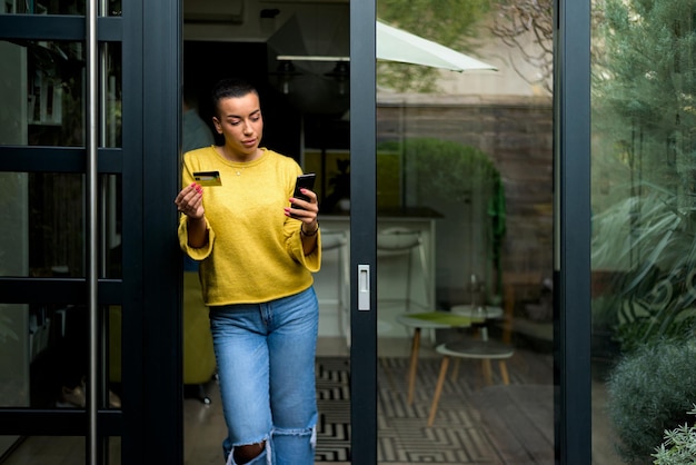 Portrait d'une femme décontractée et détendue utilisant un téléphone pour le paiement en ligne à la porte de sa maison faisant des achats en ligne depuis la maison s'appuyant sur le cadre de la porte à l'aide d'une carte de crédit entrepreneur occasionnel faisant des affaires à domicile