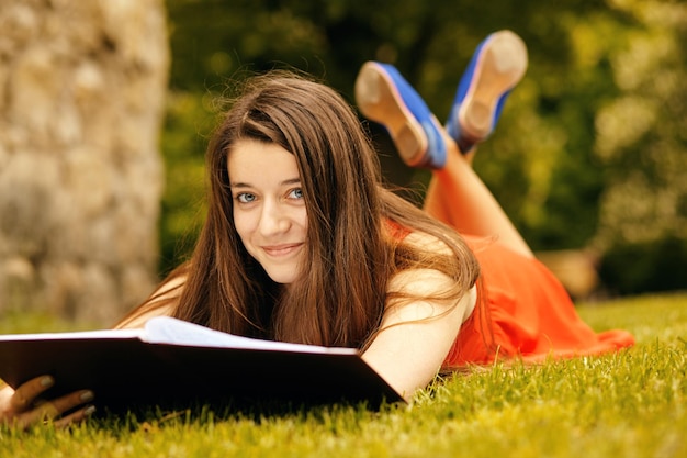 Portrait d'une femme décontractée heureuse en robe allongée sur l'herbe verte. Elle lit un livre.