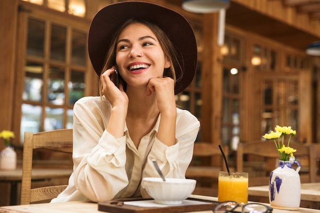 Portrait de femme détendue portant chapeau parlant sur smartphone, assis dans un café confortable