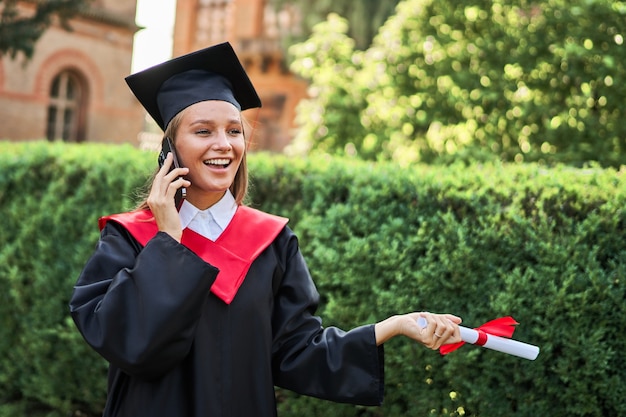 Portrait de femme diplômée souriante appelant par téléphone portable en robe de graduation avec diplôme.