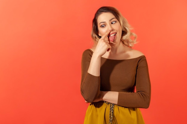 Portrait d'une femme drôle dans des vêtements décontractés touchant son nez avec l'index, les yeux croisés et la langue. Concept drôle et idiot. Tourné en studio intérieur isolé sur fond rouge