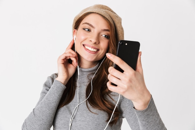 Portrait d'une femme élégante des années 30 portant des écouteurs souriant et écoutant de la musique sur un téléphone portable isolé sur blanc