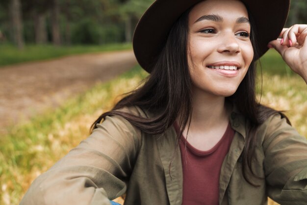 Portrait d'une femme élégante et heureuse avec de longs cheveux noirs portant un chapeau regardant de côté alors qu'il était assis sur l'herbe à l'extérieur