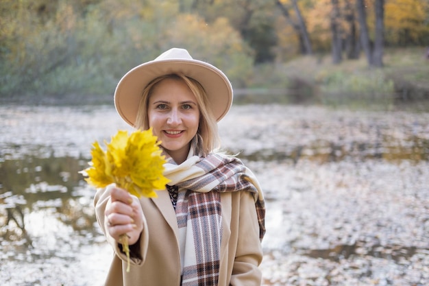 Portrait de femme élégante en manteau beige et chapeau détient des feuilles d'automne dans le parc d'automne à l'automne