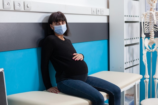 Portrait D'une Femme Enceinte Assise Sur Son Lit Dans Un Bureau De Contrôle, Attendant De Recevoir Une Consultation D'un Obstétricien. Patient Avec Masque Facial Attendant Un Enfant, Assistant à Un Rendez-vous Médical.