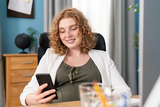 Portrait d'une femme enceinte souriante aux cheveux roux bouclés utilise un smartphone