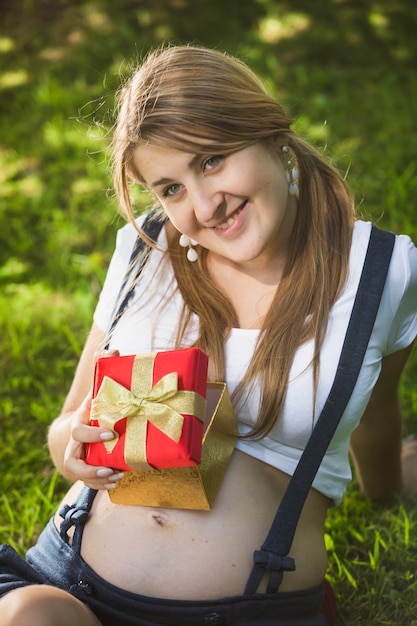 Photo portrait de femme enceinte souriante posant avec une boîte-cadeau rouge au parc