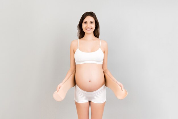 Portrait de femme enceinte en sous-vêtements habillant la ceinture de maternité contre la douleur dans le dos sur fond gris avec espace de copie. Concept de ceinture de soutien abdominale orthopédique.