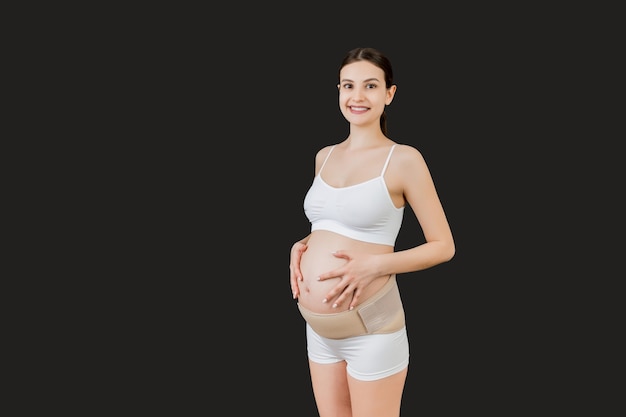 Portrait de femme enceinte en sous-vêtements portant une ceinture de maternité pour réduire la douleur dans le dos sur fond noir avec espace de copie. Concept de ceinture de soutien abdominale orthopédique.