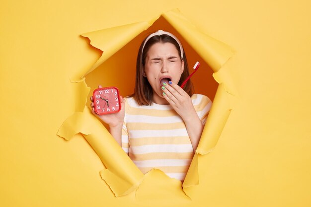 Portrait d'une femme endormie portant une chemise rayée et une bande de cheveux perce un fond de papier jaune tenant un réveil et une brosse à dents bâillant couvrant la bouche avec la paume