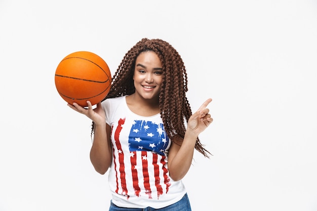 Portrait d'une femme énergique se réjouissant et tenant le basket-ball pendant le match en se tenant isolée contre un mur blanc