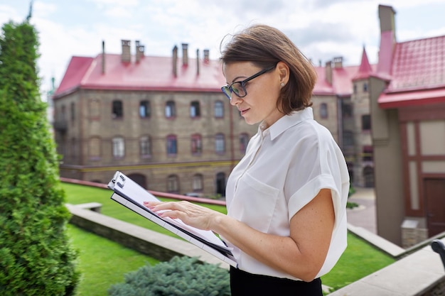 Portrait de femme enseignante avec presse-papiers, documents papier. Femme souriante de l'extérieur du bâtiment de l'école, retour à l'université collégiale