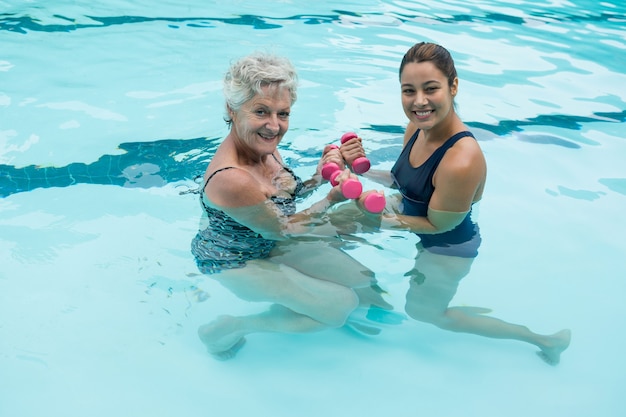 Portrait de femme entraîneur aidant senior woman à soulever des haltères dans la piscine