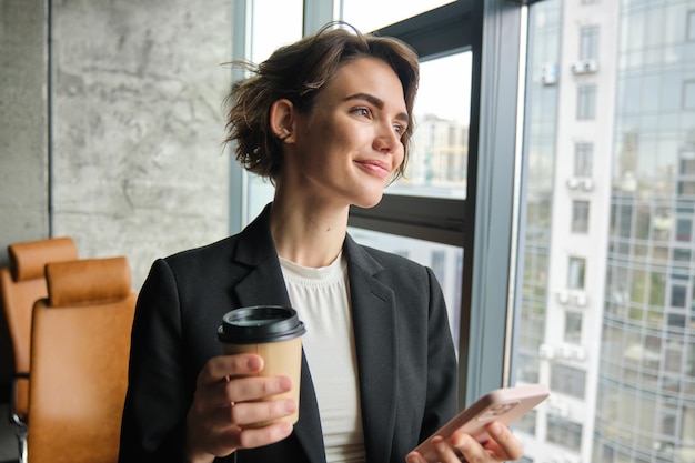 Portrait d'une femme entrepreneur à succès boit une tasse de café dans un bureau et attend un smartphone