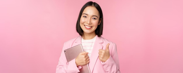 Portrait d'une femme d'entreprise fille au bureau en costume d'affaires tenant une tablette numérique montrant les pouces vers le haut recommandant une entreprise debout sur fond rose