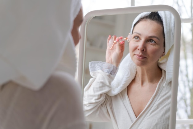 Portrait femme enveloppée dans une serviette blanche et en peignoir regarde la réflexion dans le miroir pour masser son visage avec un rouleau à billes en forme de Y Concept de cosmétiques naturels lissage des rides Mise au point sélective