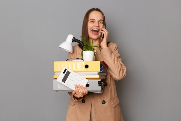 Portrait d'une femme étonnée excitée portant une veste beige tenant des dossiers en papier posant isolé sur fond gris parlant sur un téléphone portable racontant son nouveau travail tenant ses affaires