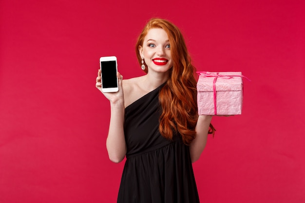 Portrait de femme excitée et amusée souriante, rousse demandée n'a pas reçu le cadeau si rapidement, montrant l'application de téléphone mobile, l'application de livraison en ligne, maintenez le rose présent sur le mur rouge