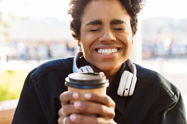 Portrait d'une femme excitée avec les yeux fermés tenant une tasse de papier en marchant dans la rue de la ville