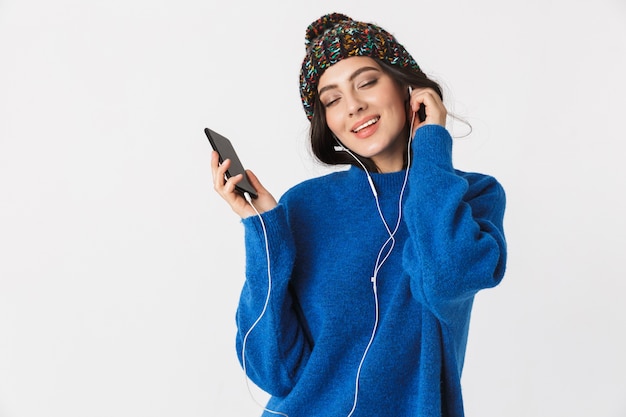 Portrait de femme extatique portant chapeau d'hiver et écouteurs tenant le smartphone en position debout, isolé sur blanc