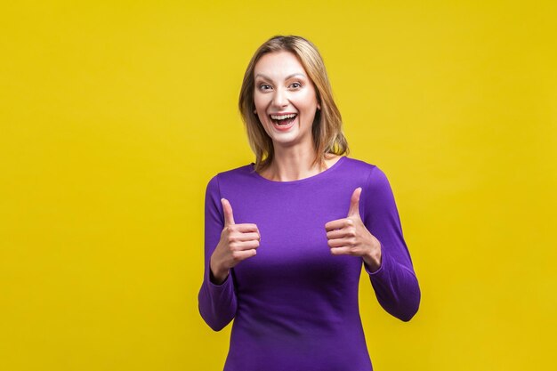 Portrait d'une femme extrêmement heureuse en robe violette serrée montrant les pouces vers le haut et souriant largement à la caméra expression excitée ravie studio intérieur tourné isolé sur fond jaune