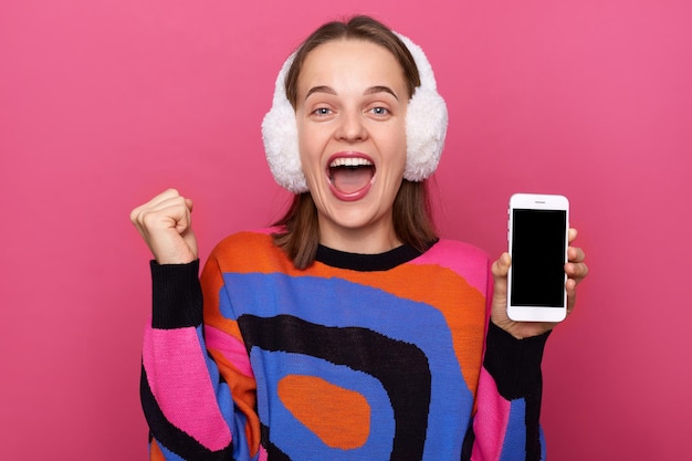 Portrait d'une femme gaie extrêmement heureuse avec le poing fermé célébrant son succès montrant un téléphone portable avec un écran vide portant un pull et des cache-oreilles en fourrure posant isolé sur fond rose