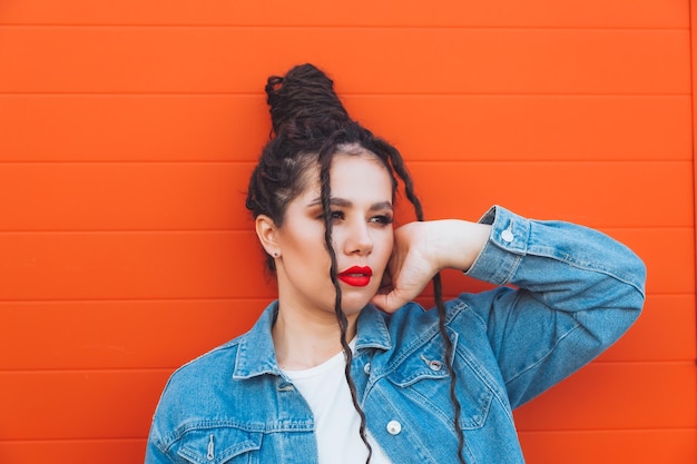 Portrait d'une femme glamour avec des dreadlocks dans un costume en jean et avec des lèvres rouges contre un mur orange