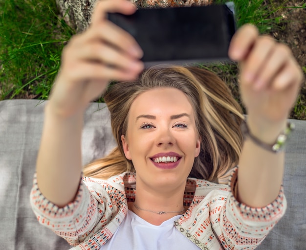 Portrait d&#39;une femme heureuse allongée dans l&#39;herbe prenant le selfie avec un téléphone intelligent. Belle femme qui se débrouille sur un gazon vert. Happy lady souriant pour la caméra et tenant un téléphone intelligent ou mobile devant elle.