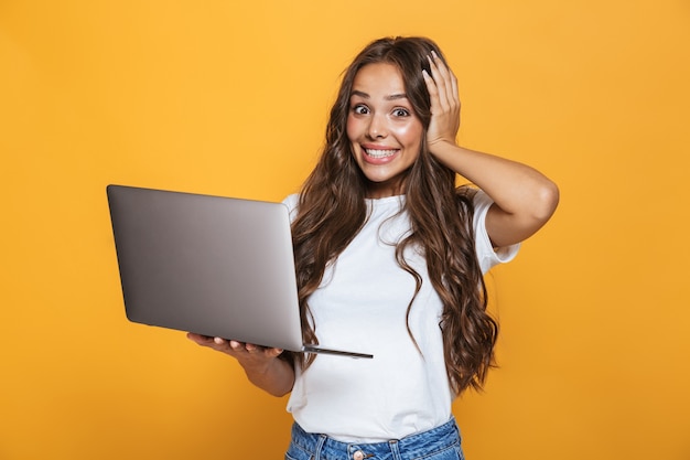 Portrait de femme heureuse des années 20 aux cheveux longs se réjouissant et tenant un ordinateur portable gris, isolé sur mur jaune