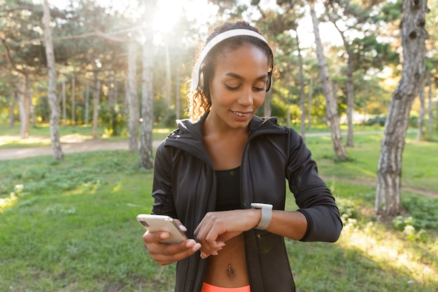 Portrait de femme heureuse des années 20 portant un survêtement noir et des écouteurs, regardant la montre-bracelet tout en marchant dans le parc verdoyant