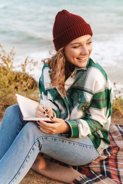 Portrait d'une femme heureuse caucasienne souriante et prenant des notes dans le journal alors qu'elle était assise sur une couverture au bord de la mer