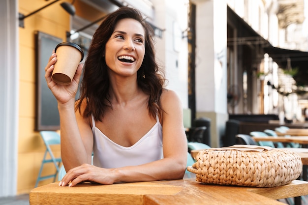 Portrait d'une femme heureuse détendue avec un sac de paille buvant du café dans une tasse en papier alors qu'elle était assise dans un café confortable à l'extérieur