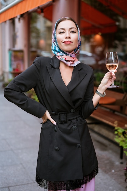 Portrait d'une femme heureuse avec du maquillage se tient près d'un café dans la rue avec un verre de vin à la main