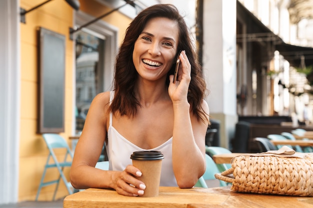 Portrait d'une femme heureuse et heureuse avec un sac de paille buvant du café dans une tasse en papier et parlant au téléphone portable tout en étant assis dans un café confortable à l'extérieur