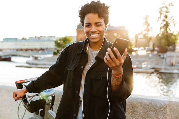 Portrait d'une femme heureuse portant des vêtements décontractés debout avec un vélo et utilisant un téléphone portable en marchant le long de la rivière