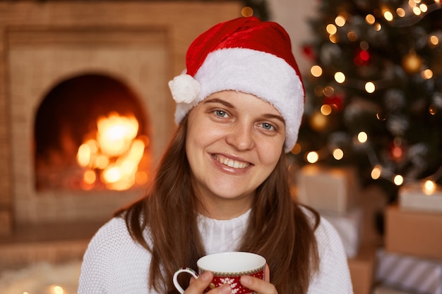 Portrait d'une femme heureuse et positive portant un pull blanc et un chapeau de père Noël, posant près d'une cheminée et d'un arbre de Noël avec une tasse dans les mains, savourant un thé ou un café chaud, regardant la caméra en souriant.