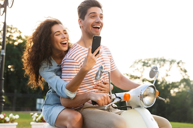 Portrait de femme heureuse prenant une photo sur smartphone, tout en roulant sur une moto à travers la rue de la ville avec son petit ami