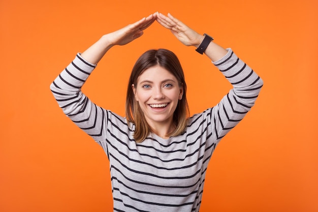 Portrait d'une femme heureuse protégée aux cheveux bruns en chemise rayée à manches longues levant les mains au-dessus de la tête faisant le toit geste assurance studio intérieur tourné isolé sur fond orange